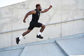 Confident strong fit sporty young black man running along urban wall. Motivated African ethnic guy jumping in air advertising sportswear, street workout training, pursuing goal concept. Side view