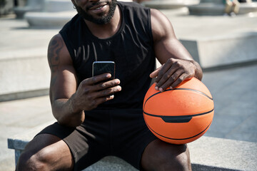 Fit sporty young African black ethnic man sitting outdoors holding basketball ball and mobile phone...