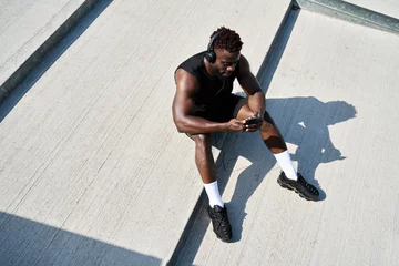 Papier Peint photo Lavable Fitness Fit sporty young black man sitting on concrete urban stairs holding phone using mobile apps listening music. Strong African ethnic guy wearing headphones looking at smartphone outdoors. Top view