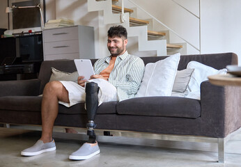 Happy young man with disability leg prosthesis sitting on sofa at home using digital tablet working or elearning, browsing web, searching online. People with amputation disabilities everyday life. - obrazy, fototapety, plakaty