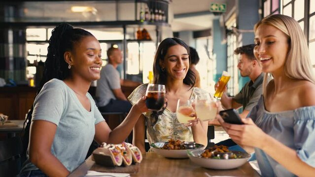 Group of multi-cultural female friends meeting in restaurant posing for selfie on mobile phone as food arrives and doing cheers  - shot in slow motion