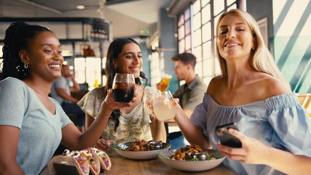 Group of multi-cultural female friends meeting in restaurant posing for selfie on mobile phone as food arrives and doing cheers  - shot in slow motion