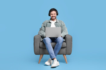 Happy man with laptop and headphones sitting in armchair on light blue background