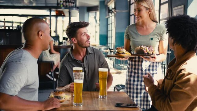 Group of multi-cultural male friends meeting in bar being served meal or burger and fries with beer by waitress - shot in slow motion