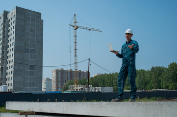 A male builder in a helmet stands against the background of a construction site with a laptop in his hands. 