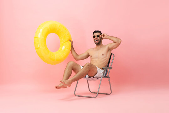 Happy Shirtless Caucasian Tourist Man Sitting On Beach Chair Holding Swim Ring In Isolated Pink Color Studio Shot Background, Summer Vocation Concept