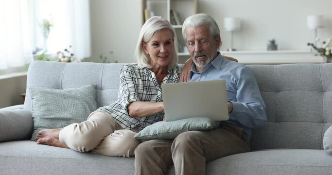 Happy aged couple spend time together at home, sit on couch use laptop, look at screen, do online shopping, choose travel offer, discuss internet news, chat with children, buy through Generative AI