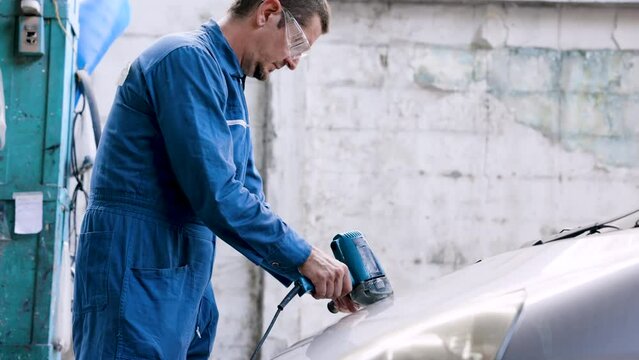 Hand male mechanic wearing suit for working in garage for safety in risky work, wear dustproof goggles, mechanic standing using polishing machine scrub surface car that comes garage make new color.