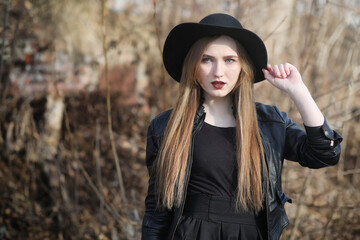 A girl walks down the city street in a leather waistcoat with a phone. Young beautiful girl in a hat and with a dark make-up outside. Girl in the Gothic style on street.