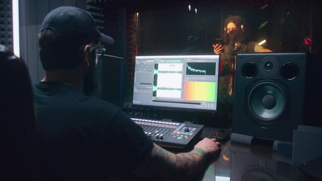 Male singer records song in soundproof room. Audio engineer, producer uses control surface. Computer screen showing DAW software interface for creating music. Sound recording studio. Music production.