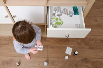 Toddler baby opened the cabinet drawer with pills and medicine. Child boy holding a pack of pills...