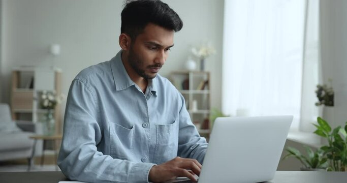 Serious Indian business man sit at desk working on computer at home office, type text, chat with colleagues, look pensive or concerned, solve business by e-mail, work on-line, do remote Generative AI