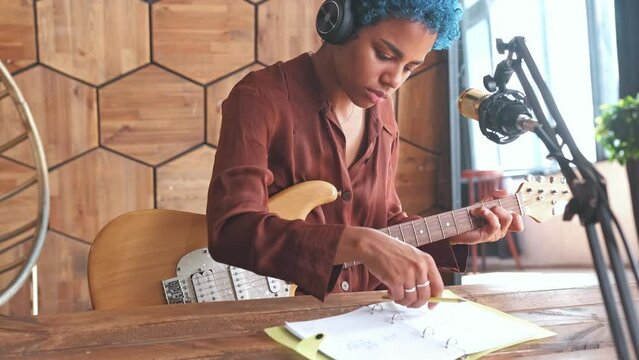 Young attractive African American woman composer with guitar invents music and writes down chords in notebook remixing popular track sits at table with microphone in recording studio.
