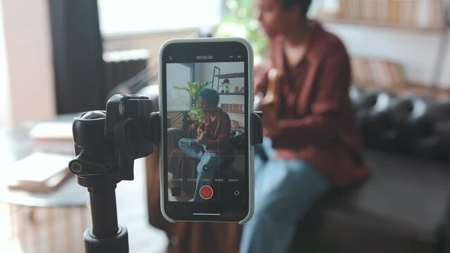 Young popular African American woman singer plays guitar and sings recording video on phone mounted on tripod to please blog subscribers with new track sits on sofa in apartment.