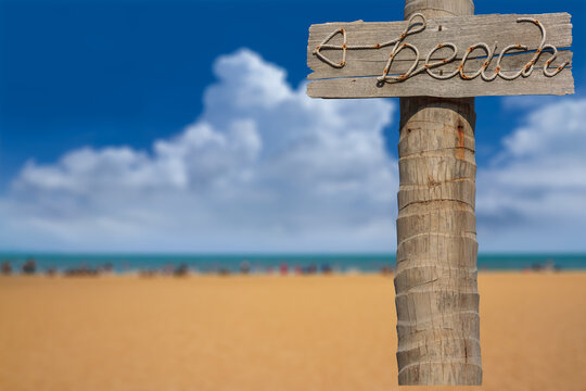 Beach sign made from manila rope on old wooden board and coconut pole with blurred the beach and clear blue sky background, Image for summer theme design with copy space and selective focus