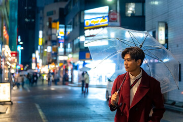 Young Asian man holding umbrella walking city street with night lights and crowd of people in Tokyo...