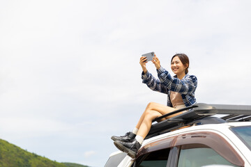Happy Asian woman sitting on car roof rack and using mobile phone taking selfie with beautiful nature. Attractive girl have fun outdoor lifestyle travel forest road trip on summer holiday vacation.