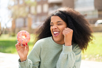 Young African American woman holding a donut at outdoors celebrating a victory
