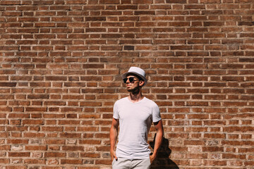 a guy with a beard in white clothes and sunglasses hold hands in pockets near the brick wall