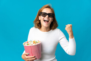 Young Georgian woman isolated on blue background with 3d glasses and holding a big bucket of popcorns