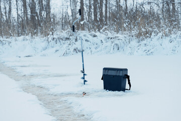 Fishing box, fishing rod and ice drill near hole on ice in winter.