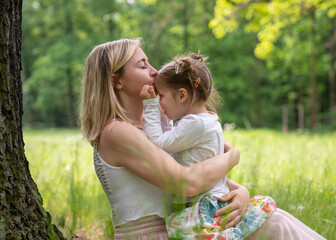 Mom and daughter in the park. Mom sits near a tree and gently kisses her little daughter. Sunny...
