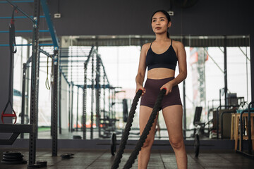 Obraz na płótnie Canvas Strong asian woman doing exercise with battle rope at crossfit gym. Athlete female wearing sportswear workout on grey gym background with weight and dumbbell equipment. Healthy lifestyle.