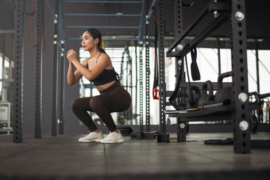 Strong Asian woman doing squat exercise at gym. Athlete female wearing sportswear workout on grey gym background with weight and dumbbell equipment. Healthy lifestyle.