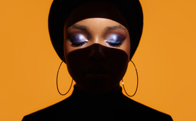 Bright make-up shadows on the eyes of an African girl are highlighted by a strip of light. Orange background.