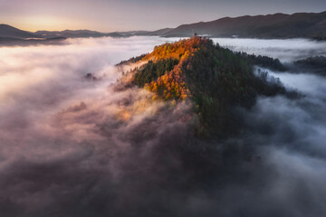 Drone view on the beautiful autumn mountains hills in low clouds at sunrise. Aerial view of Mountain peak with sunlight and colorful forest in fog. Slovenia