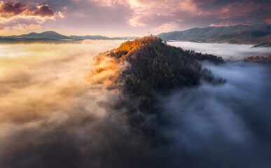 Aerial view of Mountain peak with sunlight and colorful forest in fog. Drone view on the beautiful colorful autumn mountains hills in low clouds at sunset or sunrise. Slovenia