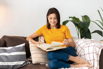 Pretty Caucasian smiling woman relaxing at home sitting on sofa reading book