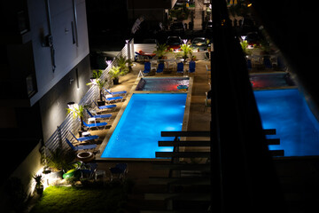 View from the balcony with the empty pool with colored lights, at night. The pool is suitable for families with children, it has deckchairs and a few palm trees around it.