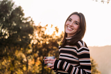 young woman smiling happy in nature looking at camera with a mug of hot drink in hand, concept of morning bliss and wilderness lifestyle, copy space for text