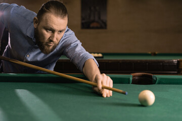 concentrated caucasian man in casual clothes playing billiards. He leaned over the table and makes a blow with a cue on the ball.