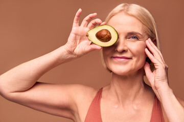 A beautiful smiling senior woman holds half an avocado covering her eye on a beige background. Beauty, health care.