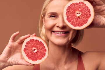 Cropped image of beautiful adult smiling blonde woman posing with grapefruit halves on beige background. Beauty, health care.