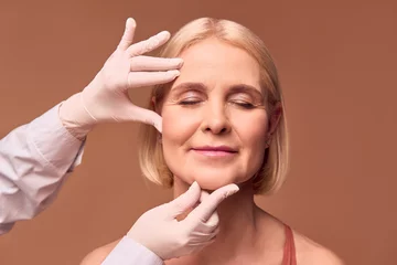Fotobehang Oude deur Portrait of an older adult woman with closed eyes on a beige background. Hands in white gloves and a medical gown show a face that has wrinkles.Face lift.