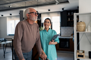 Naklejka premium Young lovely nurse helping old man to walk with walking frame. Smiling caregiver assisting elderly male at nursing home, looking at him with empathy, holding digital tablet to track recovery progress.