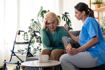 Obraz premium Worried senior woman with hand on her temple looking upset at blood pressure monitor screen concerned about high blood pressure level. Supportive nurse consoling female patient with hypertension
