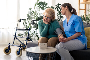 Compassionate female nurse in home visit caring for patient, showing empathy and support, sitting next to sad senior woman with hypertension. Worried senior woman concerned about high blood pressure