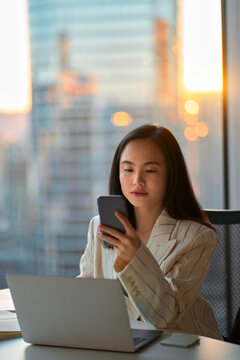 Young busy serious Asian business woman leader holding cellphone device using mobile phone, looking at smartphone checking financial market app working in modern corporate office, Generative AI