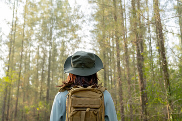 Fototapeta na wymiar Young Asian girl with a backpack and hat hiking in the mountains during the summer season, a traveler walking in the forest. Travel, adventure, and journey concept.
