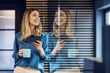 Smiling businesswoman standing in modern office with smart phone - obrazy, fototapety, plakaty