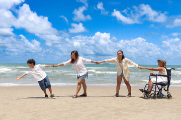 Happy disabled senior elderly woman in wheelchair spend time together with family on tropical beach. Asian grandma, daughter, grandchild boy holding hand in line, relaxing on summer holiday vacation.