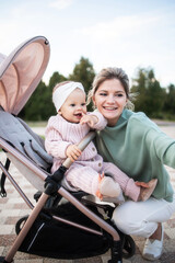 Naklejka premium Happy positive smiling young mom taking a selfie with her daughter Infant outside while walking in a stroller. Maternity leave. Maternity, Day, Mothers, Parenthood. Stylish woman, baby in stroller