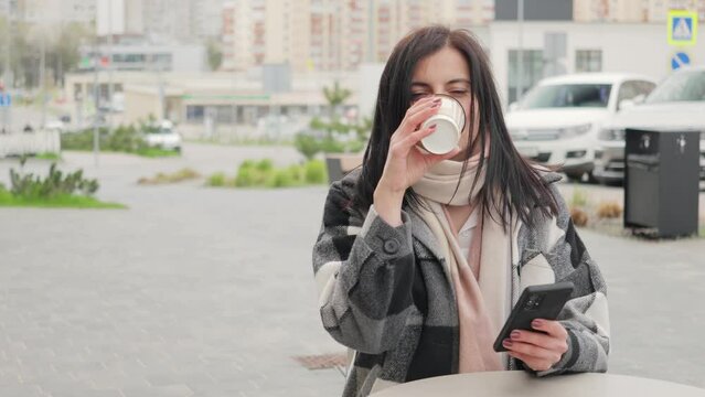 Attractive and pleasant young woman sitting at the table in a street cafe, drinking coffee from a paper cup and using smartphone for internet browsing and social media