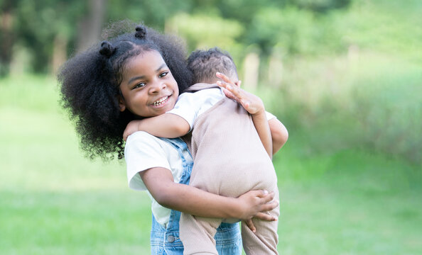 Little Cute African Older Sister With Afro Hair Smiling Carrying Her Small Brother With Good Care At Park. Sibling Relationship And Family Bond Concept