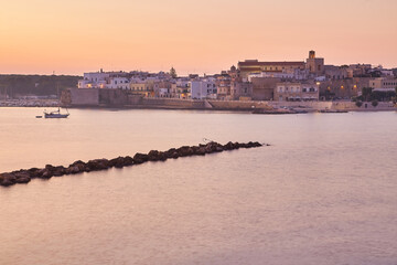 Fototapeta premium beautiful orange sunset or sunrise over the harbor of Otranto, Italy. Soft clouds, calm ocean and backlit skyline and marina with yachts and fishermen.