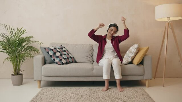 Cheerful indian woman in white headphones sitting on sofa and enjoying music Generative AI